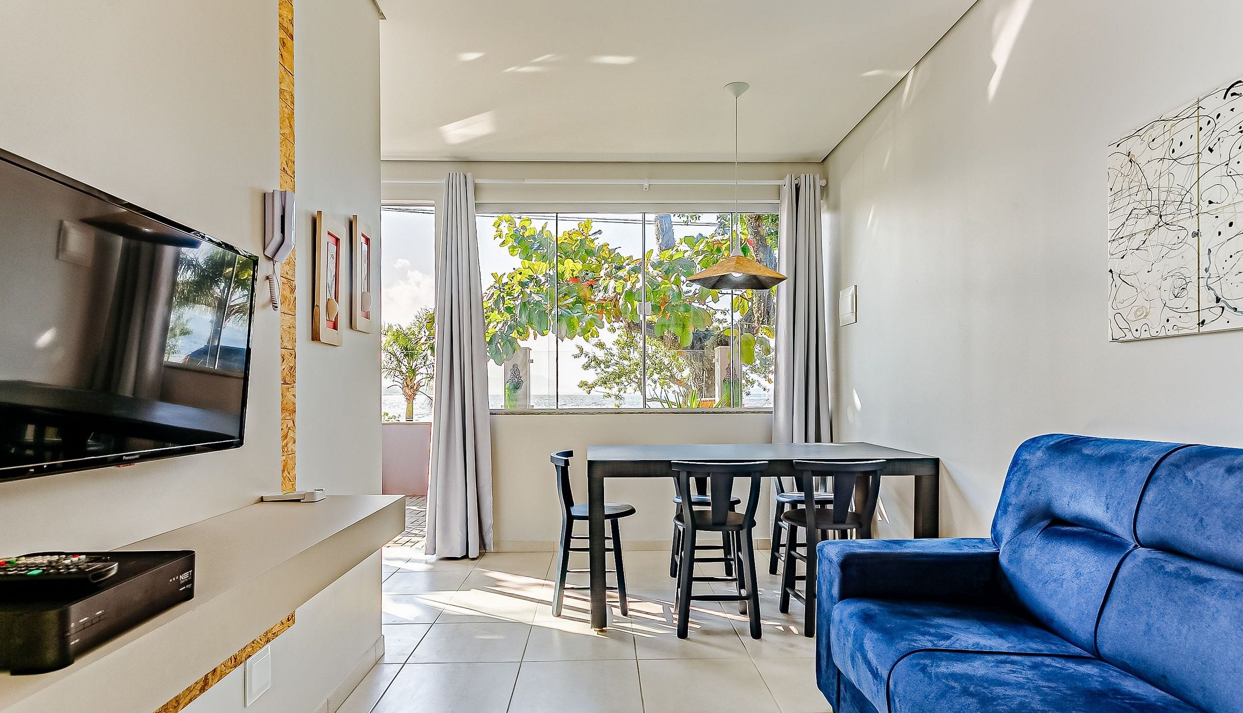 Una sala de estar moderna con un televisor de pantalla plana, un sofá azul, una mesa de comedor con sillas y un ventanal con cortinas.