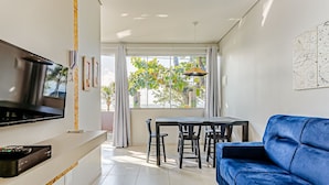 Uma sala de estar moderna com TV de tela plana, um sofá azul, uma mesa de jantar com cadeiras e uma grande janela com cortinas.