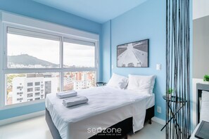 A modern hotel room with a large window, a bed with white linens, a black metal partition, and a city view. - Comfort Near Ufsc Floripa Sve407 (Florianópolis)