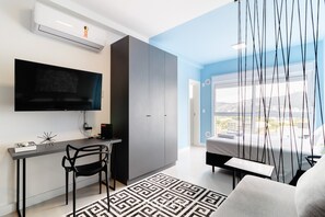 A modern room with a TV mounted on the wall, a desk with a chair, a wardrobe, a bed, and a view of the city through large windows. - Comfort Near Ufsc Floripa Sve407 (Florianópolis)