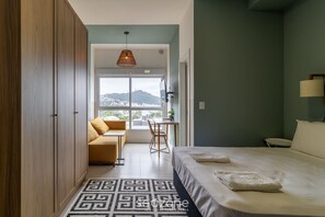 A hotel room with a bed, a desk, a chair, and a view of a cityscape through a large window. - Comfortable Studio Near the Center, Floripa Sve508 (Florianópolis)