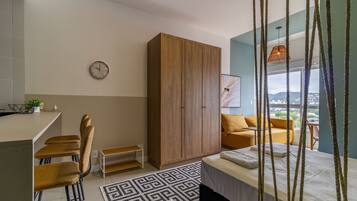 A modern bedroom with a bed, a wooden wardrobe, a dining area with chairs, a small table, and a view of the city through large windows.