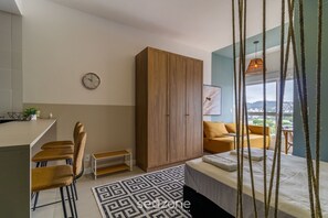 A modern bedroom with a bed, a wooden wardrobe, a dining area with chairs, a small table, and a view of the city through large windows.