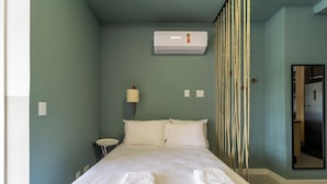 Un dormitorio con una cama, una lámpara de noche, un aire acondicionado y una puerta que da a la cocina.