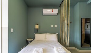 Un dormitorio con una cama, una lámpara de noche, un aire acondicionado y una puerta que da a la cocina.