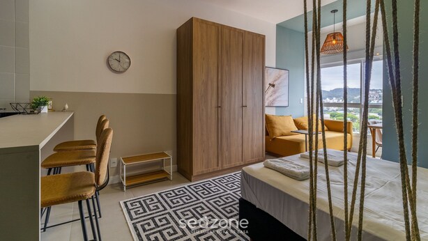 Un dormitorio moderno con cama, un armario de madera, una zona de comedor con sillas, una mesita y vistas a la ciudad a través de amplios ventanales.