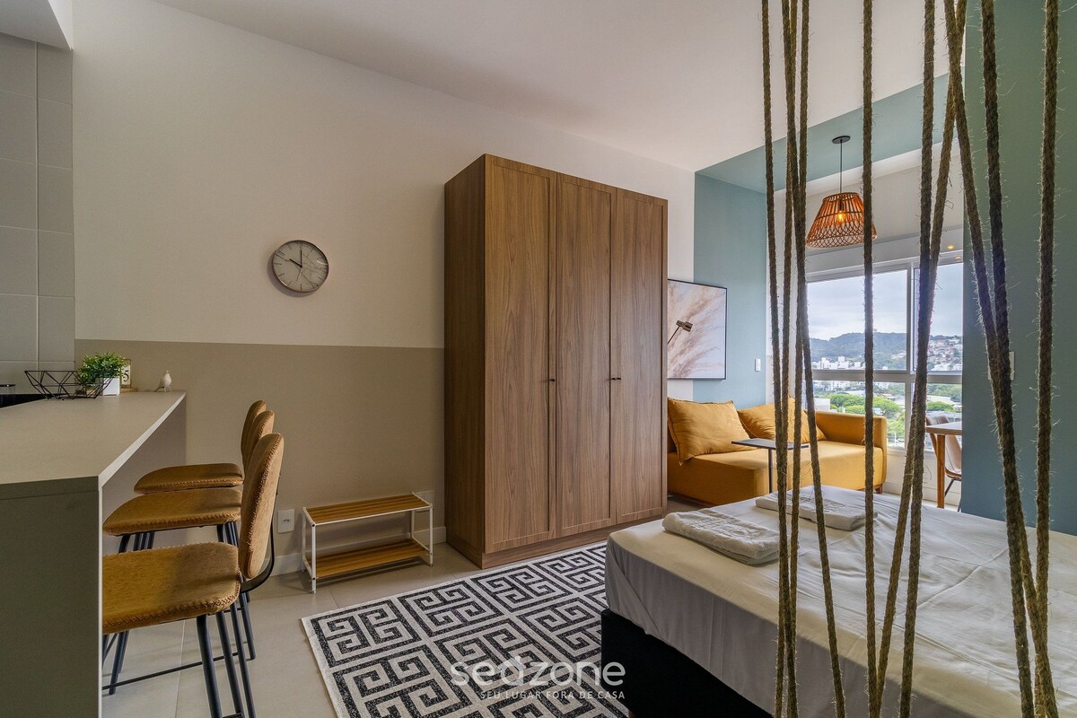 A modern bedroom with a bed, a wooden wardrobe, a dining area with chairs, a small table, and a view of the city through large windows.