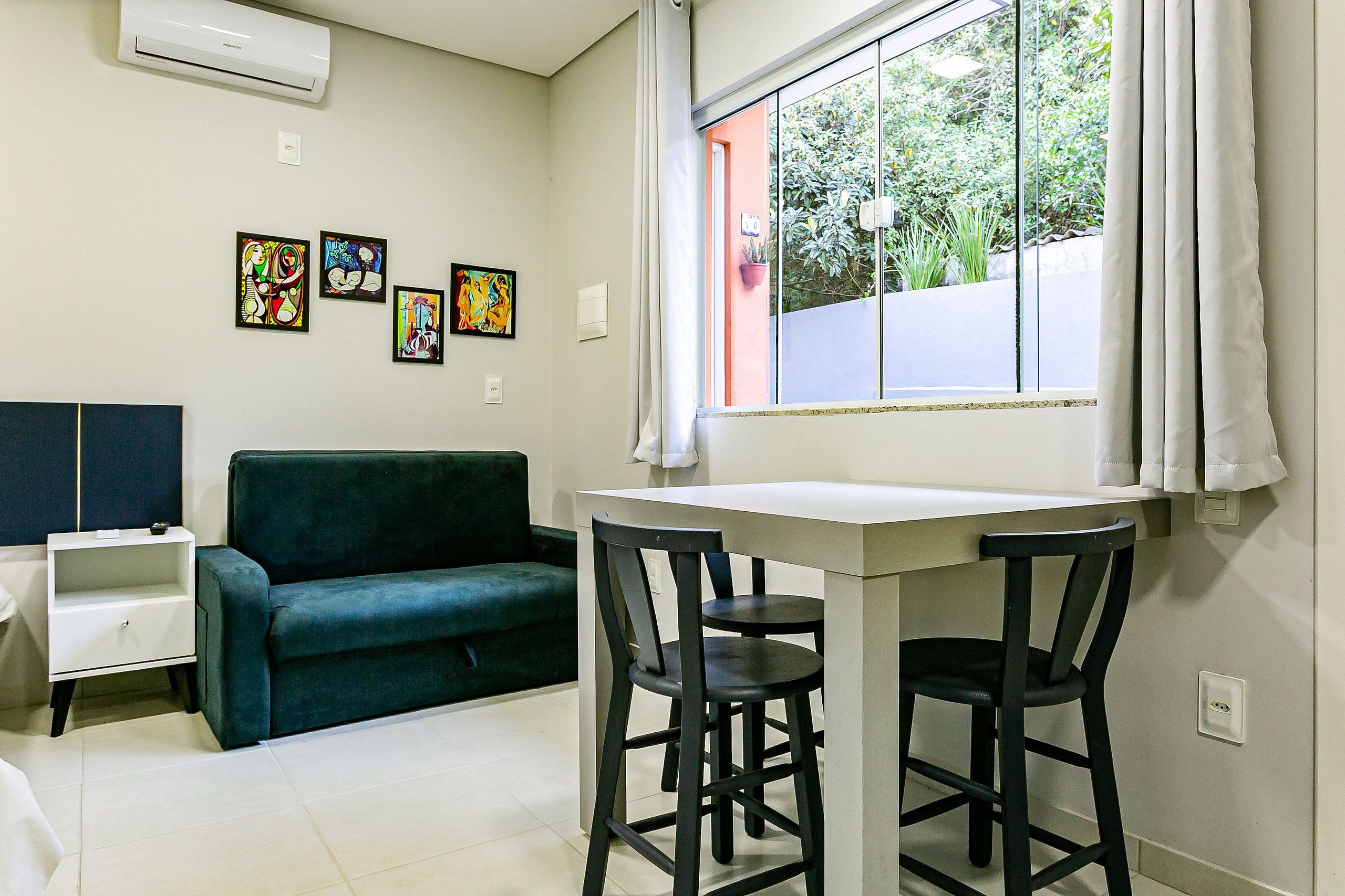 Una habitación con un sofá verde, una mesita blanca, dos sillas negras y una ventana con cortinas.