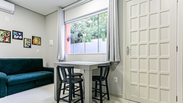 Una habitación con un sofá verde azulado, una mesa blanca con dos sillas, un ventanal con vistas a zonas verdes y una puerta blanca.