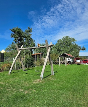 Un ensemble de balançoires en bois installé dans une zone herbeuse, avec un véhicule rouge et une petite structure en bois en arrière-plan.