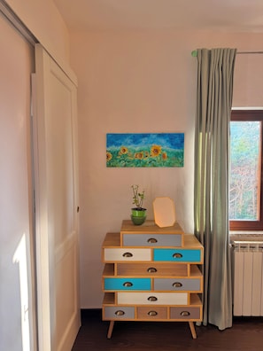 A room with a wooden dresser, a painting of sunflowers, a potted plant, and a window with curtains. - Casa Collazzi | Historic Center & Panoramic View (Montepulciano)