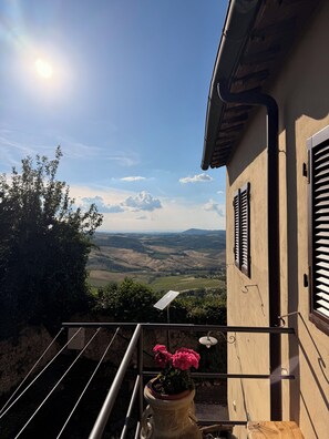 A balcony with a view of rolling hills, a potted plant with flowers, and a metal railing. - Casa Collazzi | Historic Center & Panoramic View (Montepulciano)