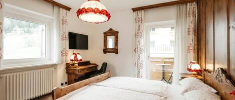 A bedroom with a large bed, a wooden desk, a chair, a television, and a window with curtains.