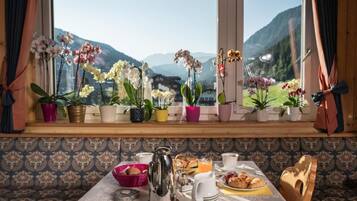Ein Frühstückstisch mit Bergblick durch ein Fenster, Zimmerpflanzen und eine hölzerne Bank.