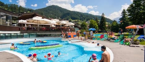 A swimming pool area with people swimming and playing, surrounded by lounge chairs and umbrellas.