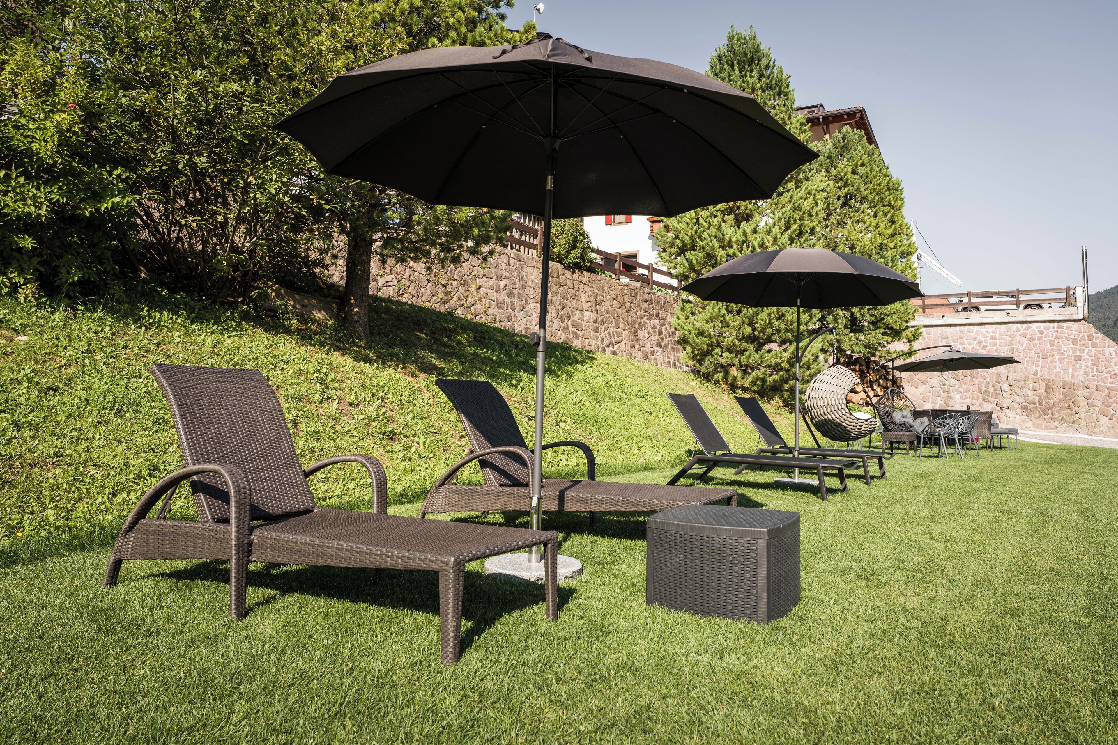 Des fauteuils de jardin et des parasols sont disposés sur une pente herbeuse, avec un mur de pierre et des arbres en arrière-plan.