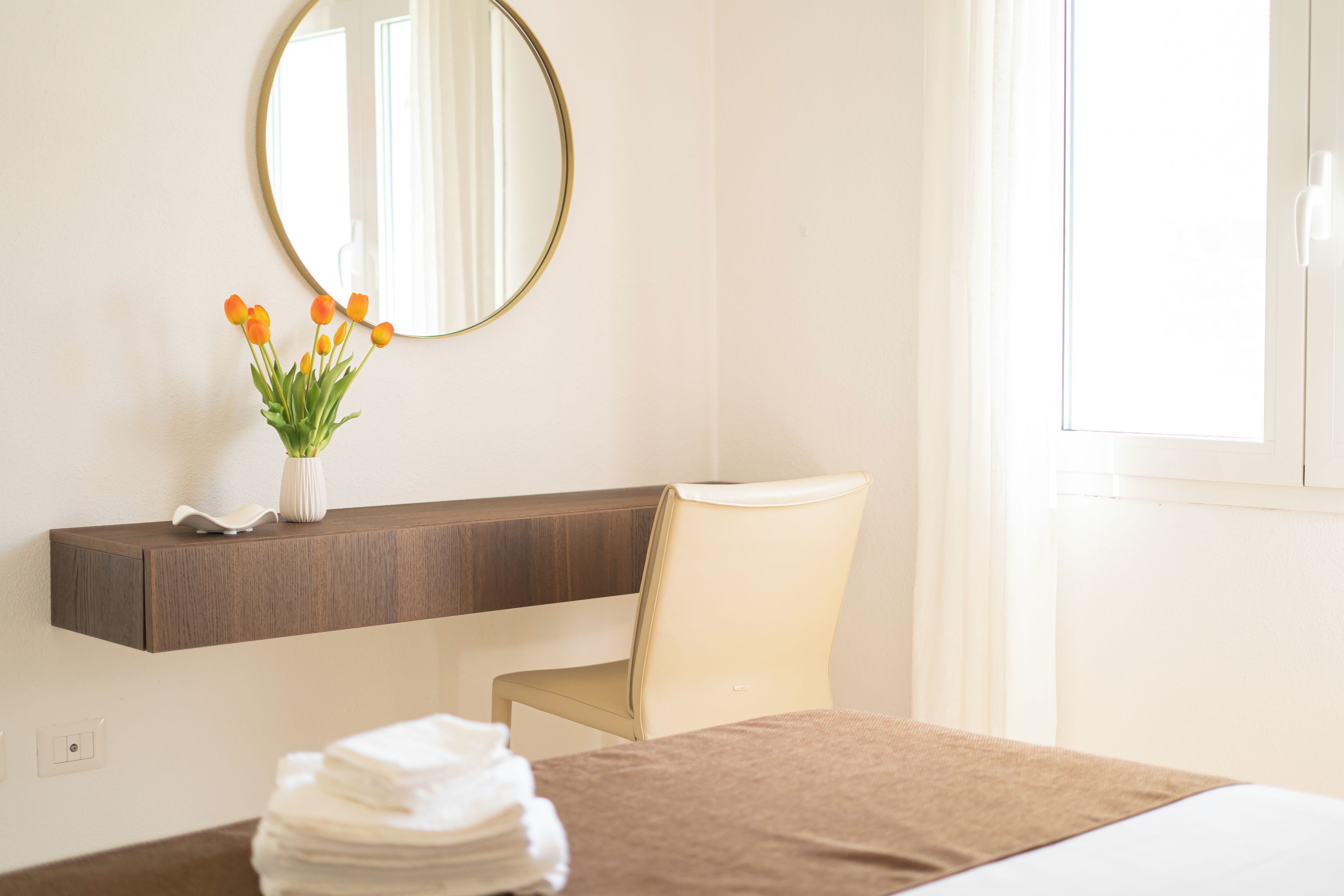 Une chambre d’hôtel avec un lit, une chaise, une table de chevet ornée d’un vase de fleurs et un miroir au mur.
