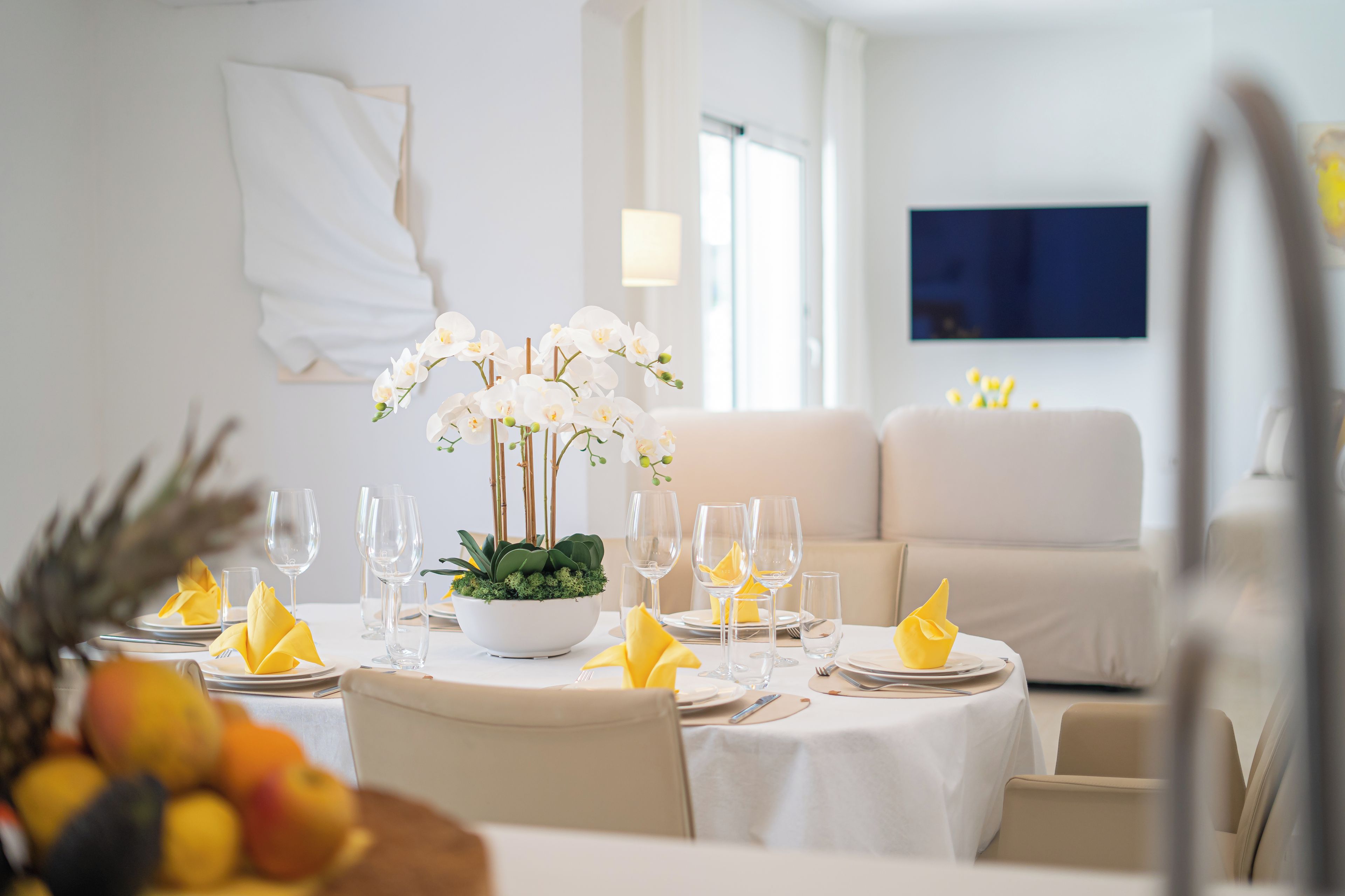 Ein Essbereich mit einem Tisch, der für vier Personen gedeckt ist, mit weißen Tischdecken, gelben Servietten und einem Mittelstück aus weißen Orchideen.