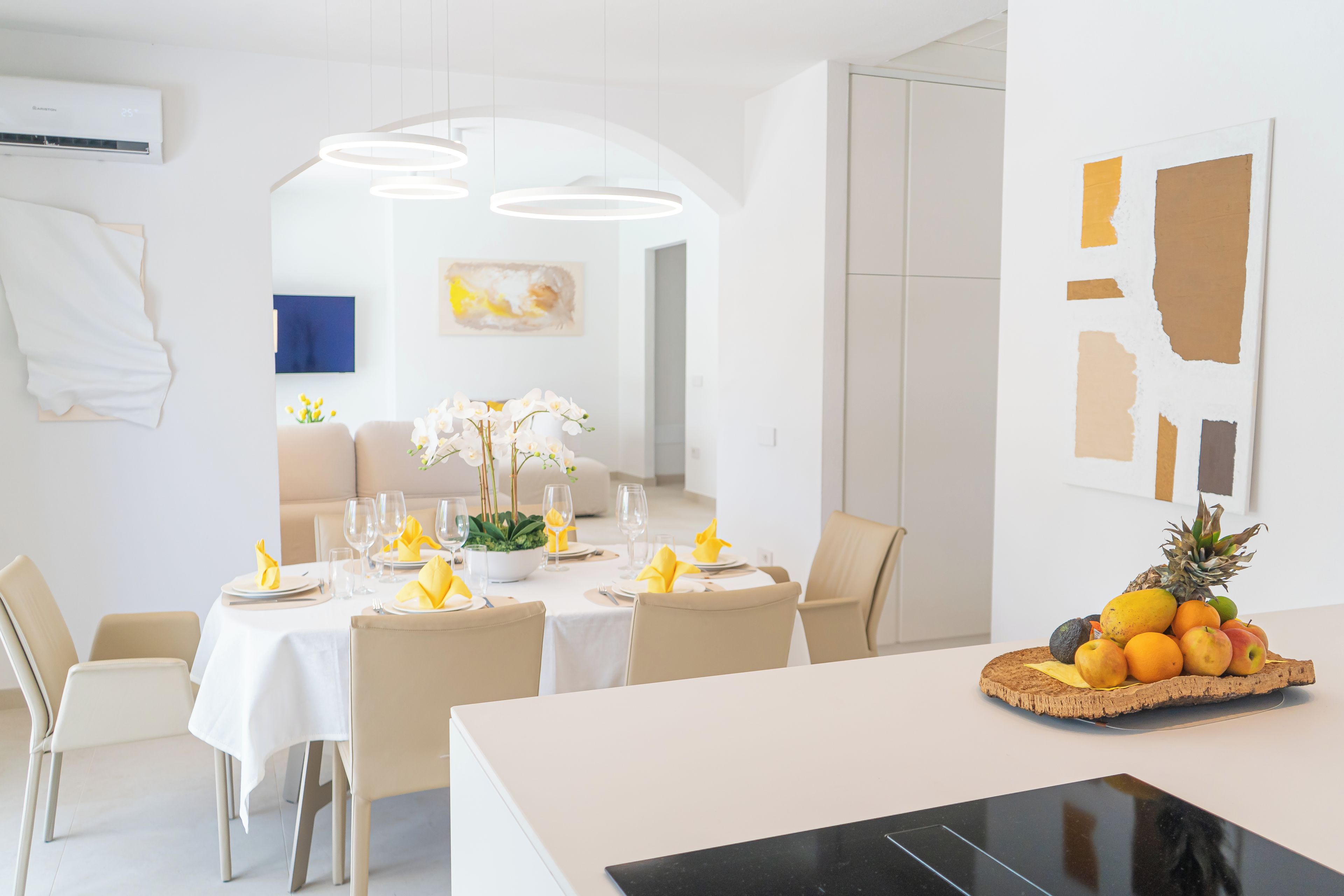 A modern dining area with a white table set for four, a fruit bowl on the table, and a painting on the wall.