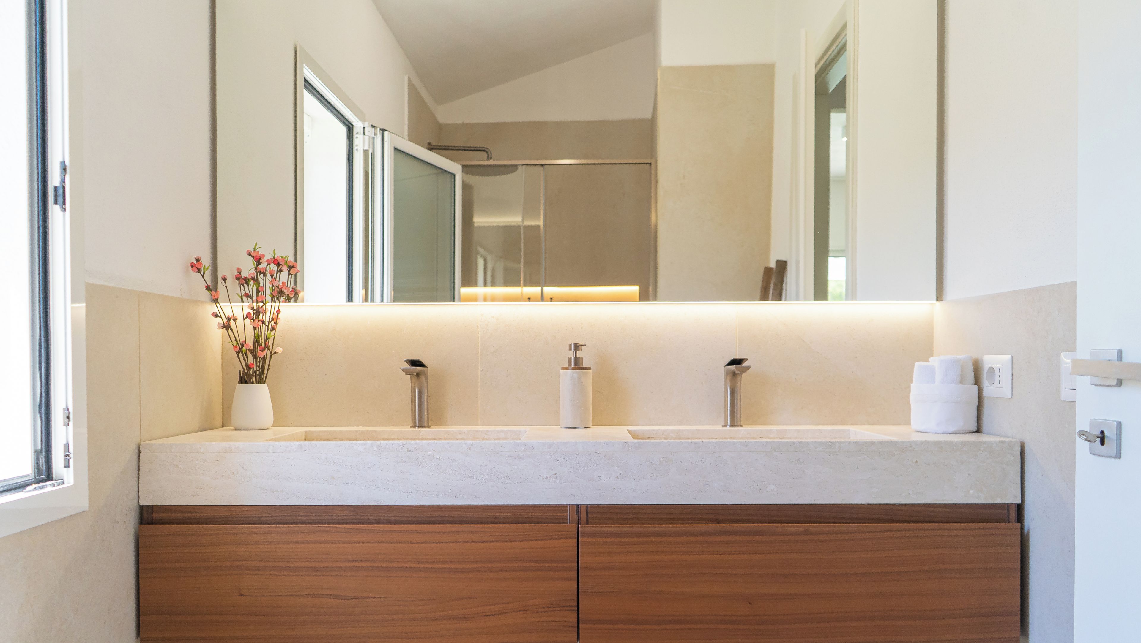A modern bathroom with a double sink, wooden cabinets, a mirror, and a vase with flowers.