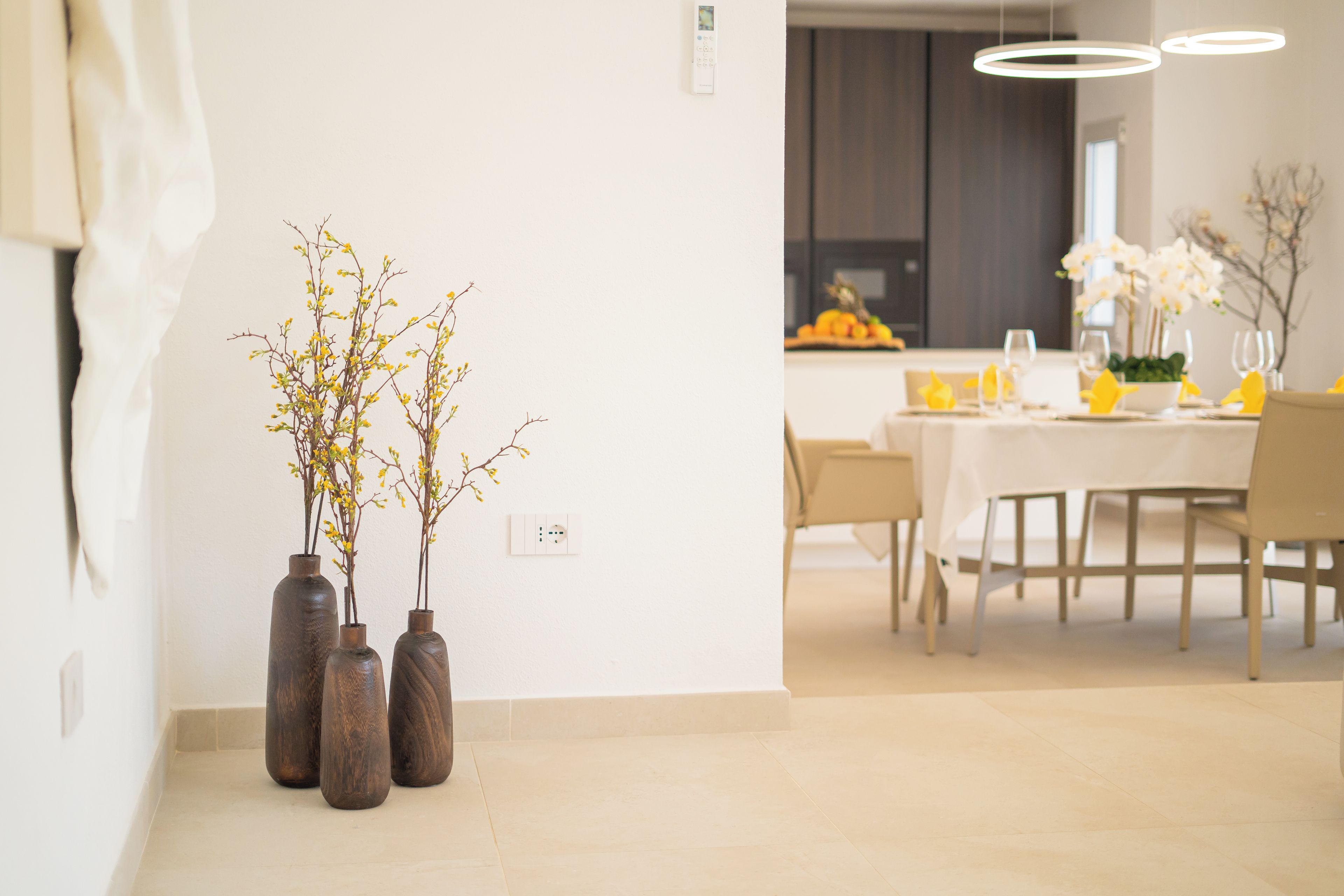 A dining area with a table set for four, a vase with branches on the wall, and a modern kitchen in the background.