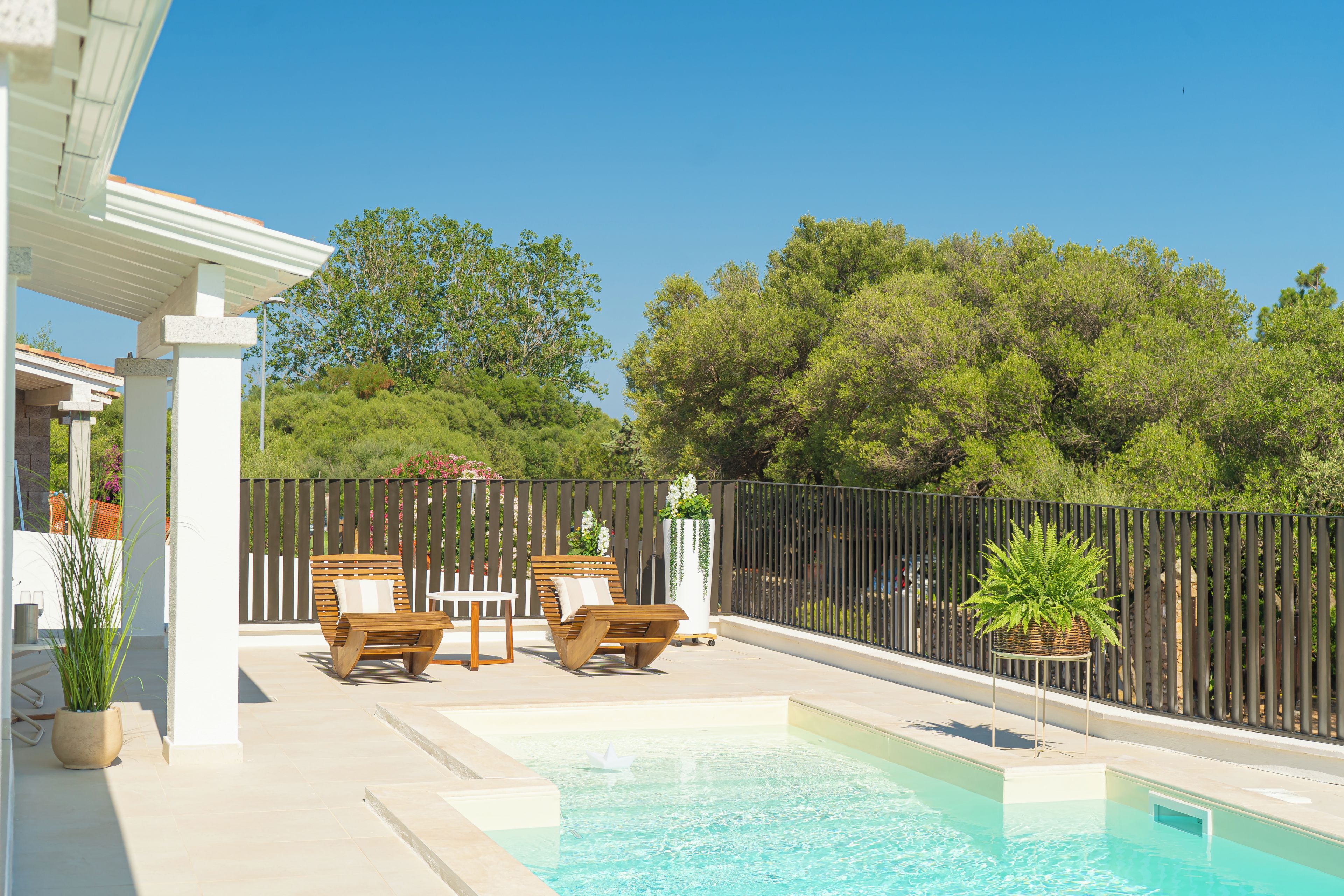 A pool area with a pergola, two wooden lounge chairs, and a small table.