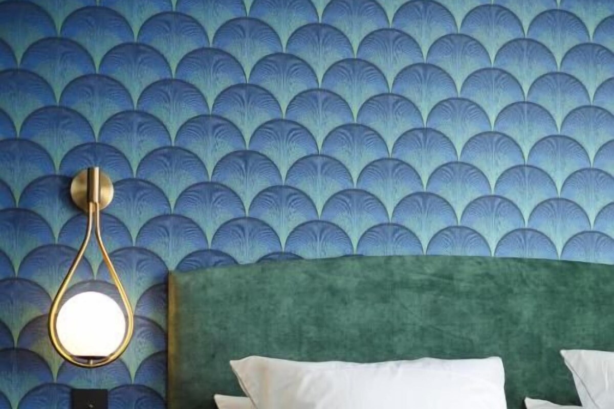 Una cama con ropa blanca, un cabecero verde y una pared con papel tapiz estampado en azul.
