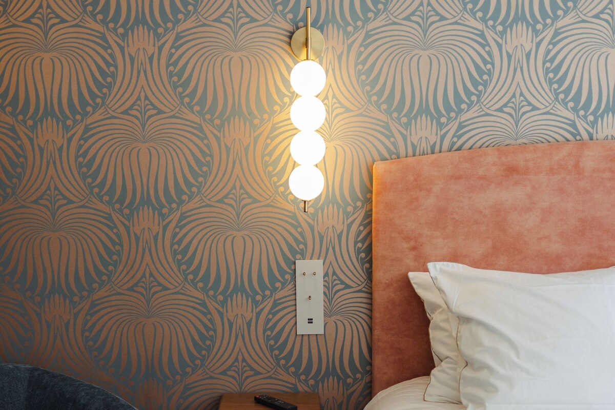 Ein Schlafzimmer mit gemustertem Wandpapier, einem Nachttisch mit Fernbedienung und einem Beistelltisch mit Lampe.