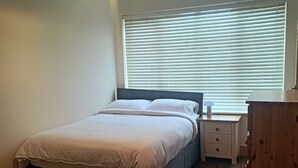 Ein Schlafzimmer mit Bett, Nachttisch, Lampe, Fenster mit Jalousien und einem Holzschrank.