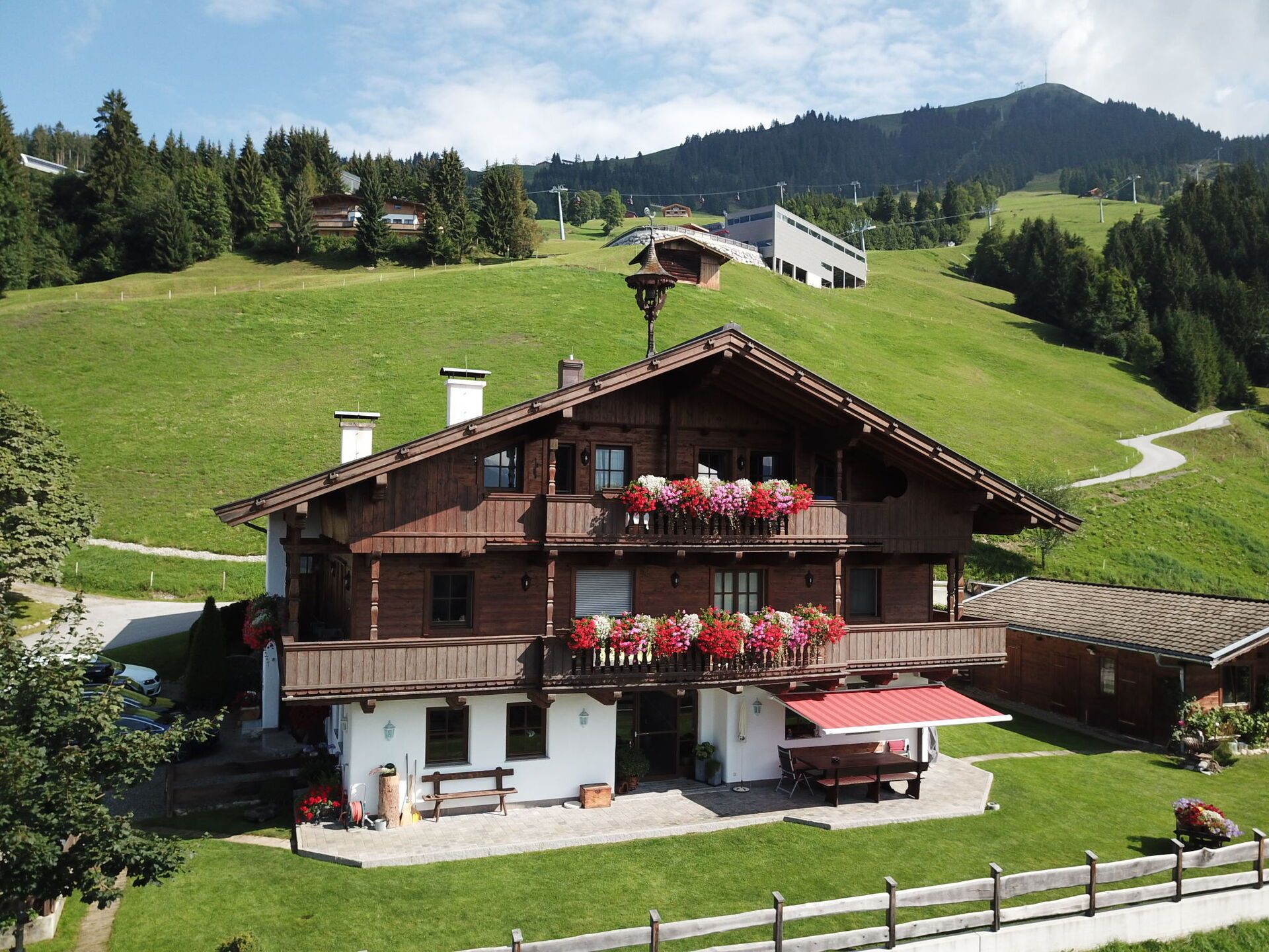 Un chalet en bois à deux étages, doté d’un balcon, entouré d’un paysage verdoyant luxuriant et bordé d’une route sinueuse.