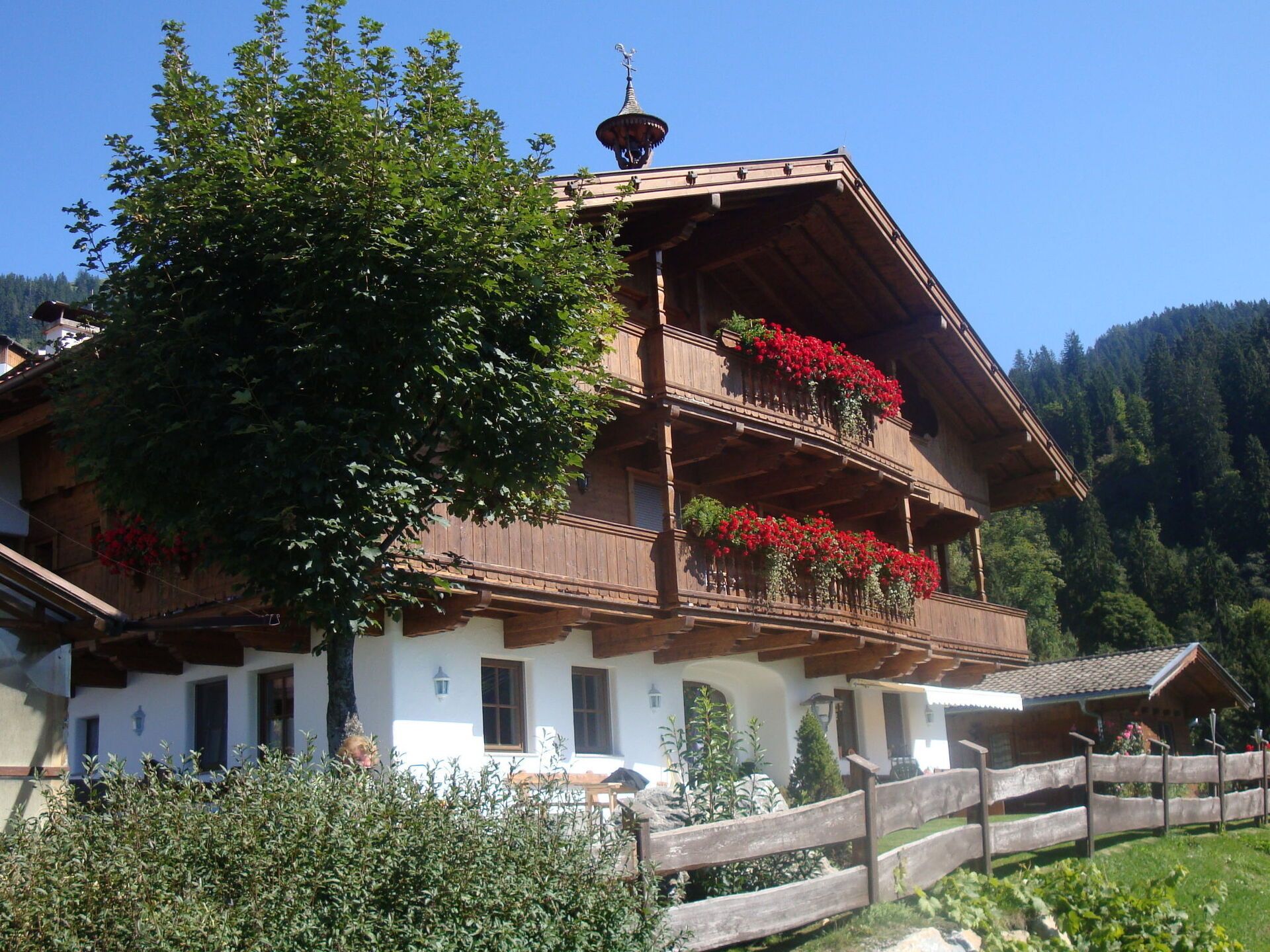 A traditional wooden chalet with a balcony adorned with red flowers, set against a mountainous backdrop.