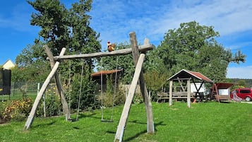 Un columpio de madera en un área cubierta de césped, con un vehículo rojo y una pequeña construcción de madera al fondo.