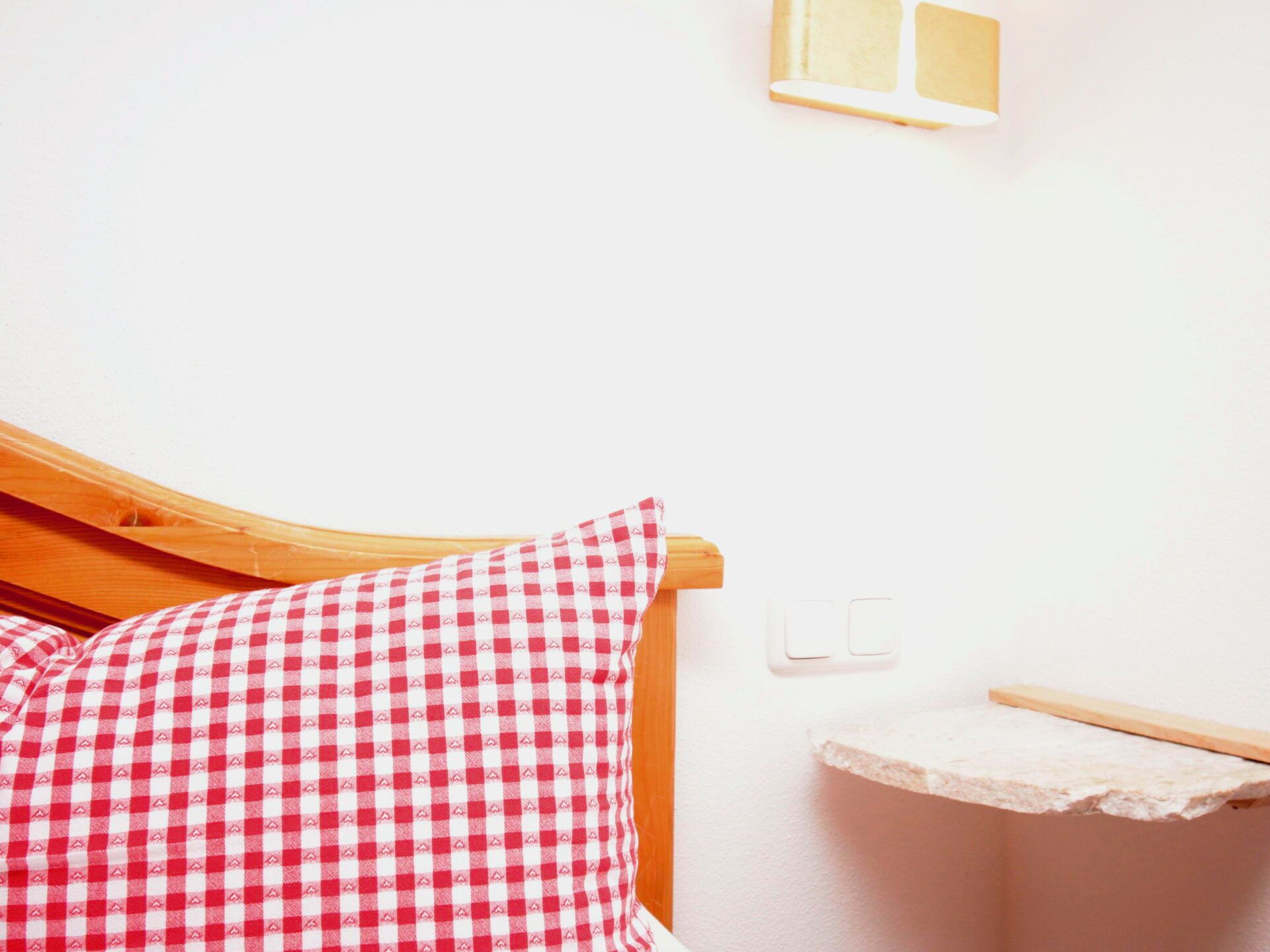 Ein Bett mit hölzernem Kopfteil, einem rot-weiß karierten Kissen und einem kleinen Nachttisch mit weißer Tischplatte.