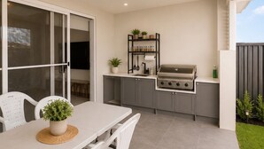 A modern outdoor kitchen with a built-in grill, a dining table with chairs, and a small garden area. - Luxury Family Retreat in Chipping Norton (Chipping Norton)