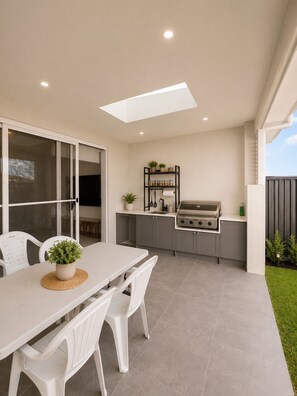 Cozinha externa moderna com churrasqueira embutida, mesa de jantar com cadeiras e um pequeno jardim.