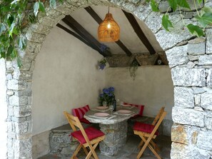 Ein von Steingewölben geschützter Sitzbereich im Freien mit einem für zwei Personen gedeckten Tisch, umgeben von Holzstühlen mit roten Kissen.