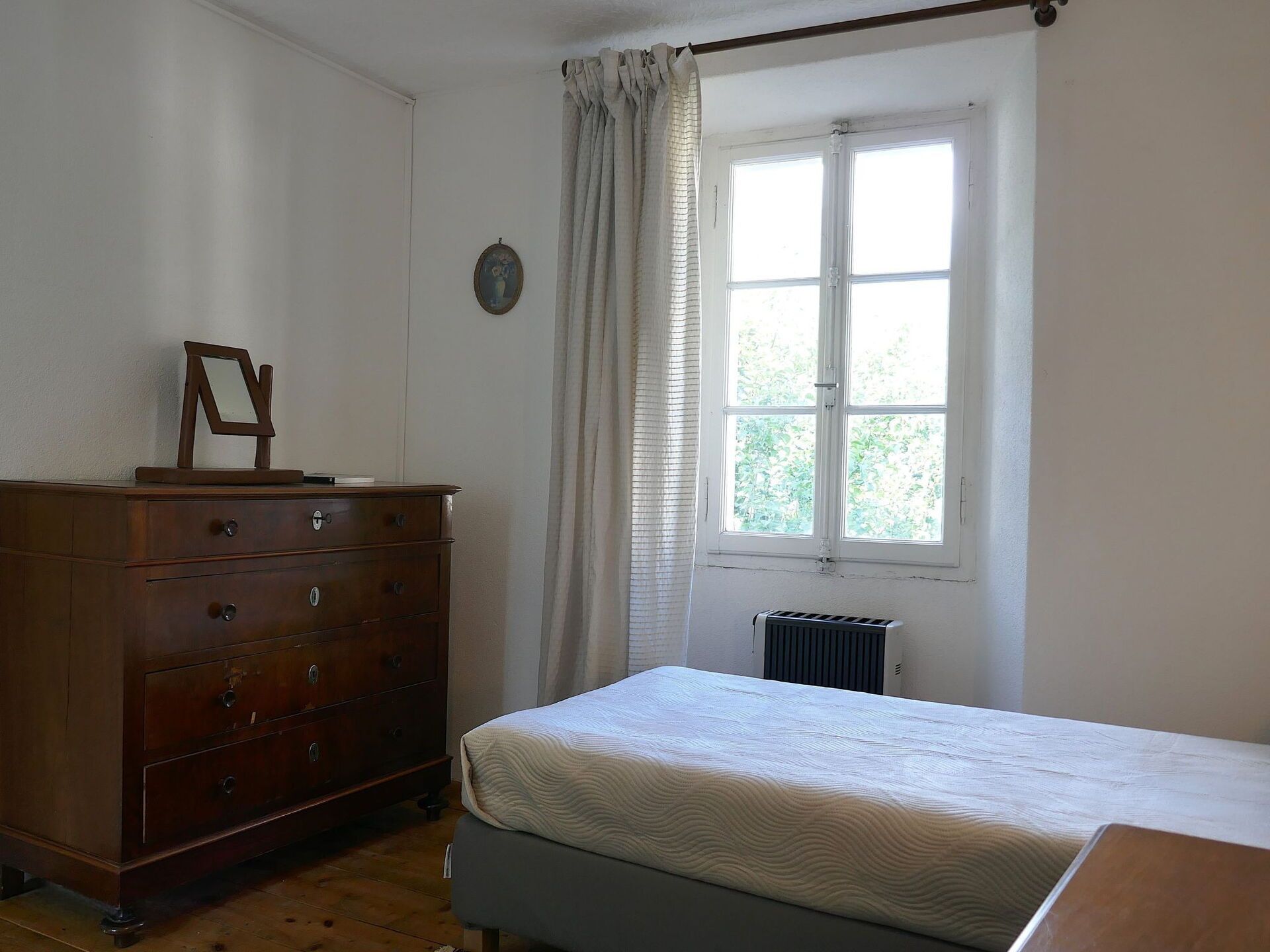 A bedroom with a wooden dresser, a bed, a window with curtains, and a radiator.