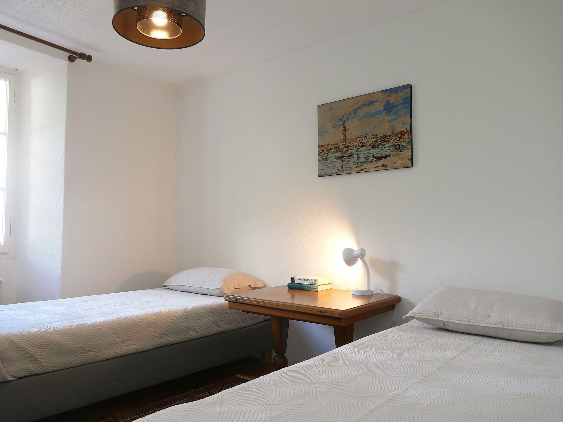 A bedroom with two single beds, a bedside table with a lamp, and a painting on the wall.