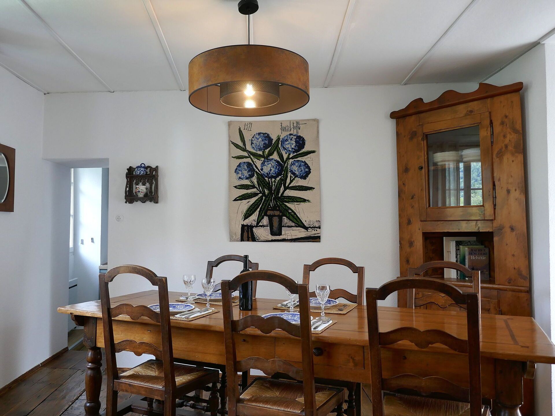Uma área de refeições com uma mesa de madeira posta para quatro pessoas, rodeada por cadeiras, um candeeiro pendente e um quadro na parede.
