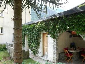 Un'area salotto esterna rustica con muri in pietra e una porta in legno, immersa nel verde con le montagne sullo sfondo.