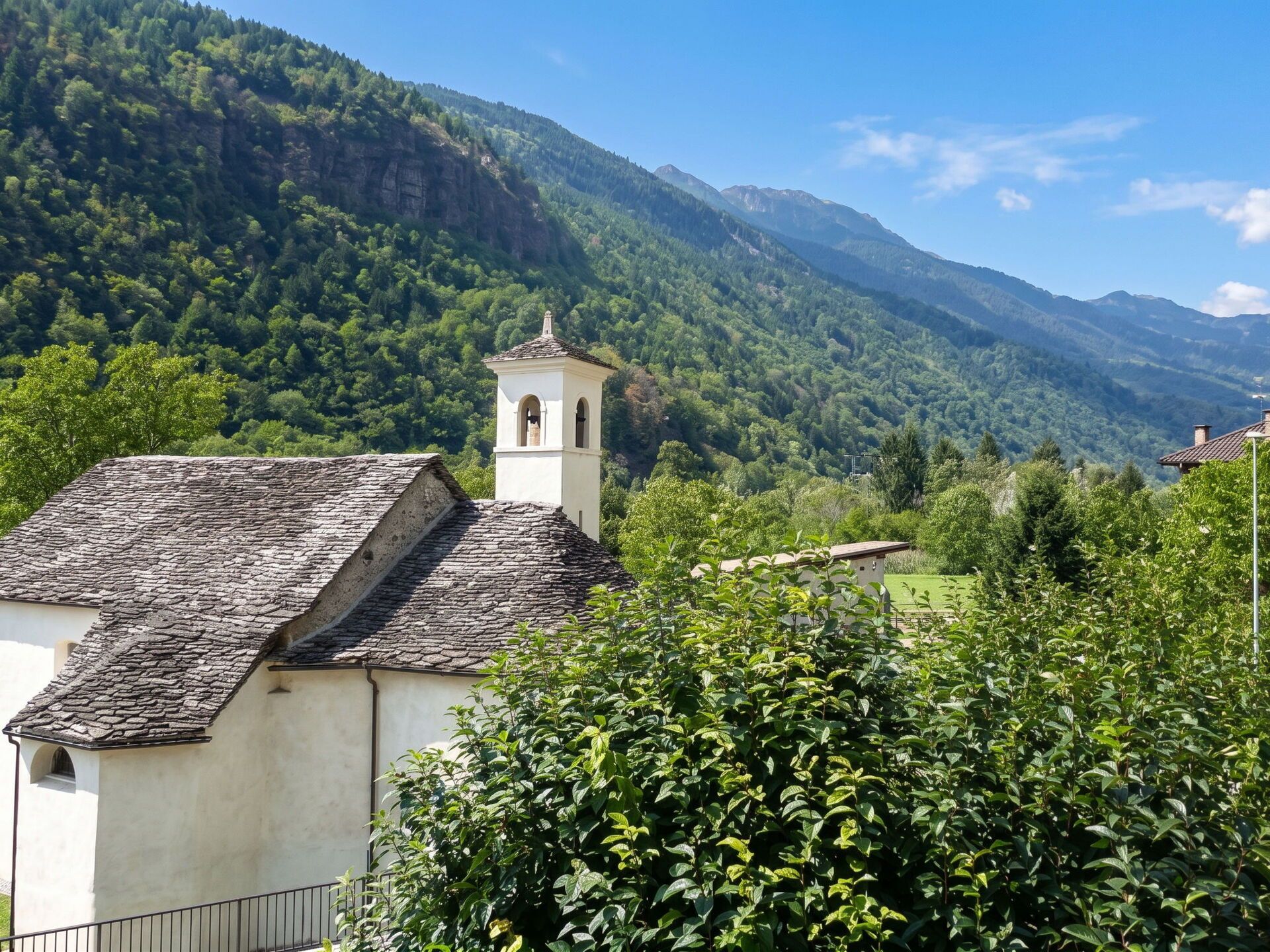 Um edifício rústico com telhado de pedra, rodeado por vegetação exuberante e montanhas ao fundo.