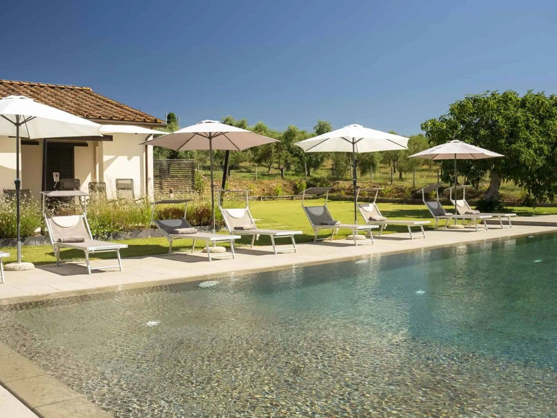 Un espace aménagé au bord de la piscine, avec des chaises longues et des parasols, une piscine à eau claire et un bâtiment doté d’un toit en tuiles.