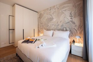 Een hotelkamer met een groot bed, een nachtkastje met lamp, een kledingkast en een muur met een tropisch schilderij.