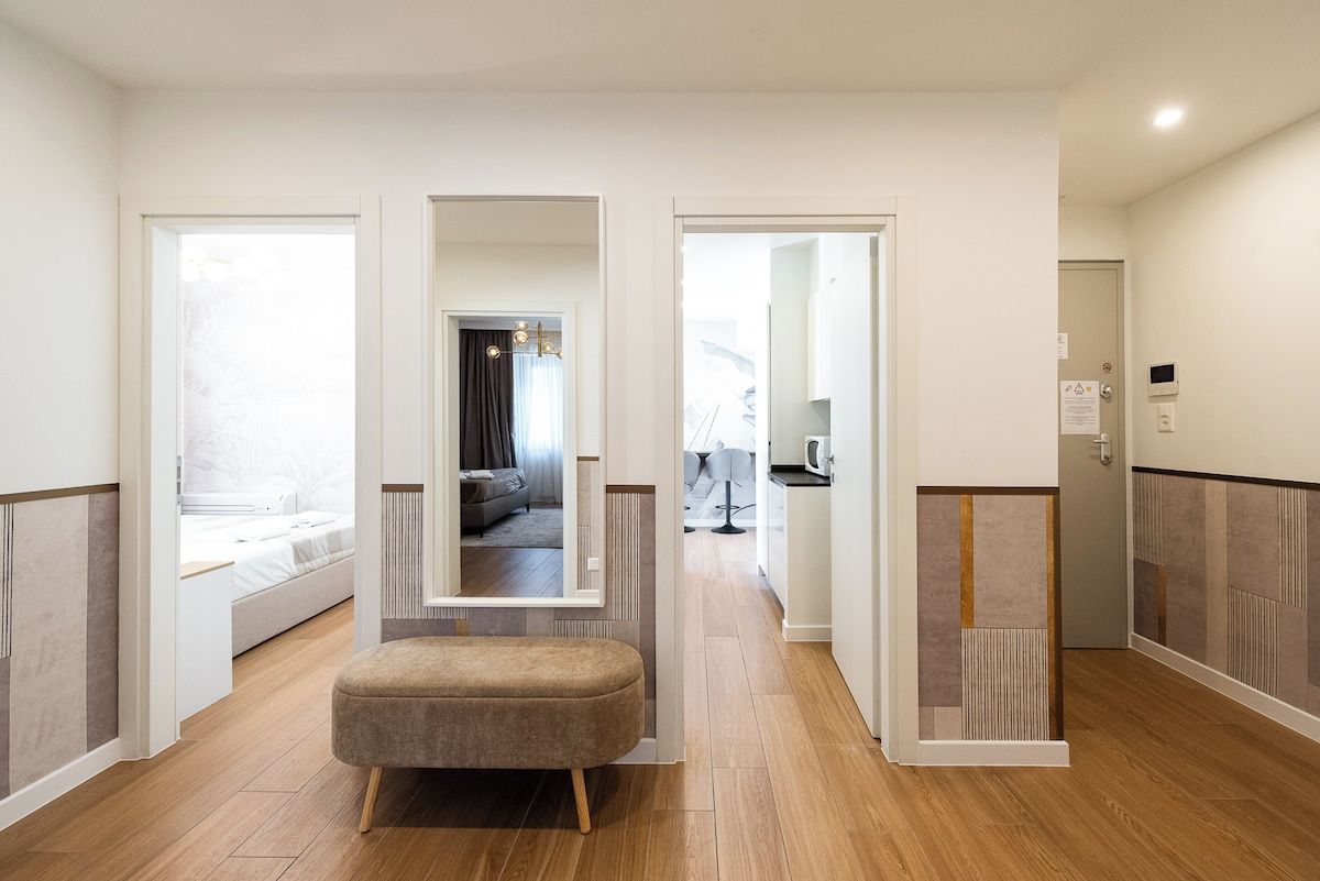 A modern hotel room with a wooden floor, a bed, a seating area, and a view into the living area.