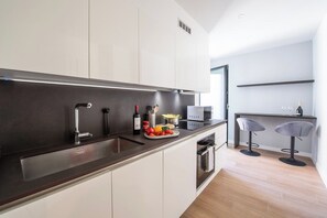 A modern kitchen with a dark countertop, white cabinets, a built-in oven, and a microwave. There are two bar stools and a small bar area with a bottle and glasses. - Boutique Suites Two Bedroom Apartment with AC (Lugano)