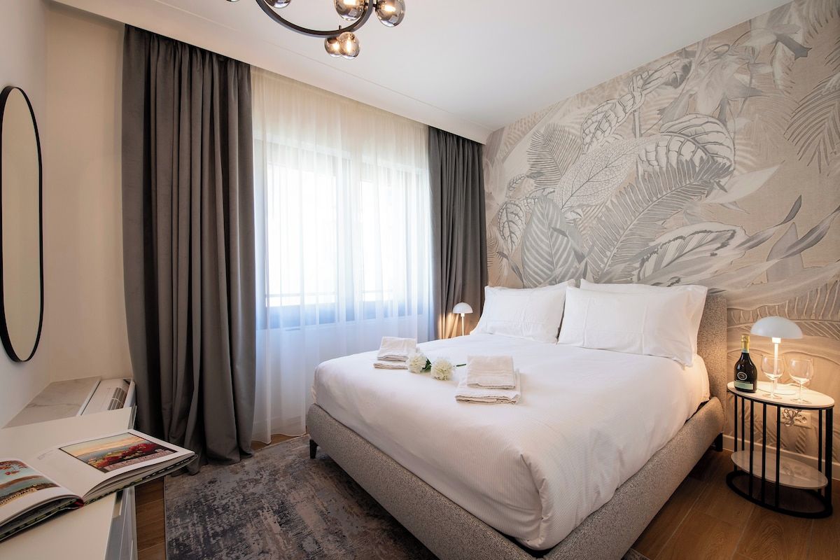 Habitación de hotel con una cama grande, una mesita de noche con lámpara, una ventana con cortinas y una pared con diseño de hojas.