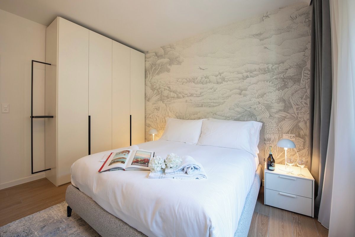 A bedroom with a large bed, a nightstand with a lamp, a wardrobe, and a wall with a nature-themed mural.