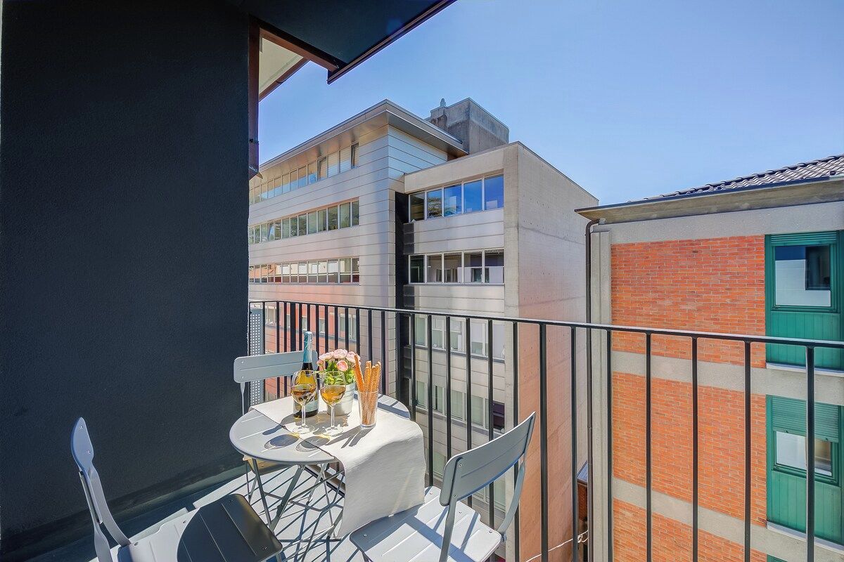 Un balcon avec une table dressée pour deux personnes, une chaise et une vue sur les bâtiments voisins.