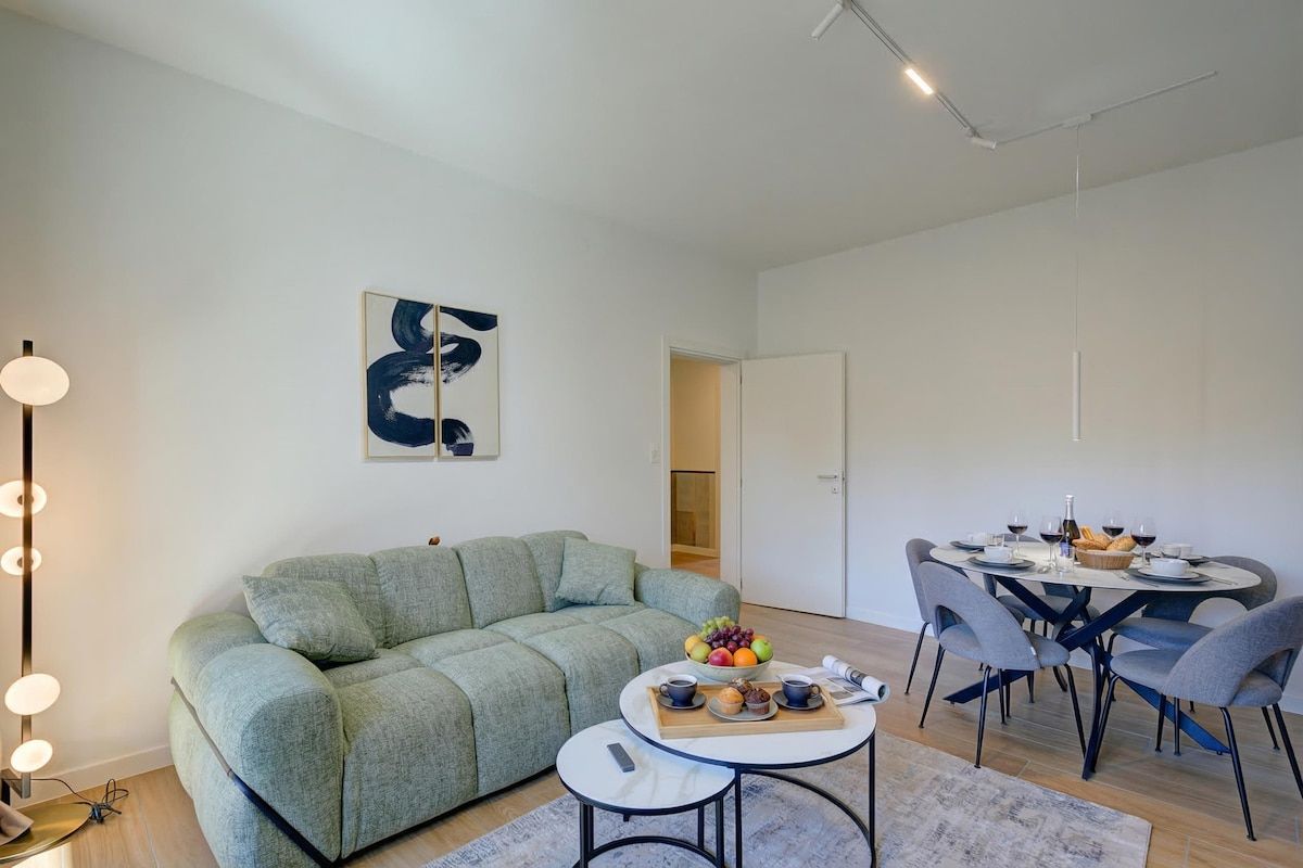 A modern living room with a green sofa, a round dining table with chairs, and a coffee table with a fruit bowl and cups.