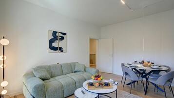 A modern living room with a green sofa, a round dining table with chairs, and a coffee table with a fruit bowl and cups.